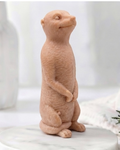 A highly detailed handmade vegan soap, sculpted in the shape of a realistic meerkat standing upright in its iconic sentinel pose, featuring textured fur and a curious facial expression, displayed on a light wooden surface.