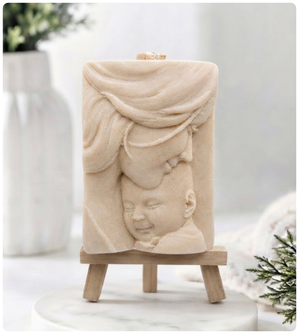 A rectangular handmade vegan soap featuring a high-relief carving of a mother tenderly cradling a small baby in her arms, displayed on a miniature wooden easel in a bright farmhouse setting.