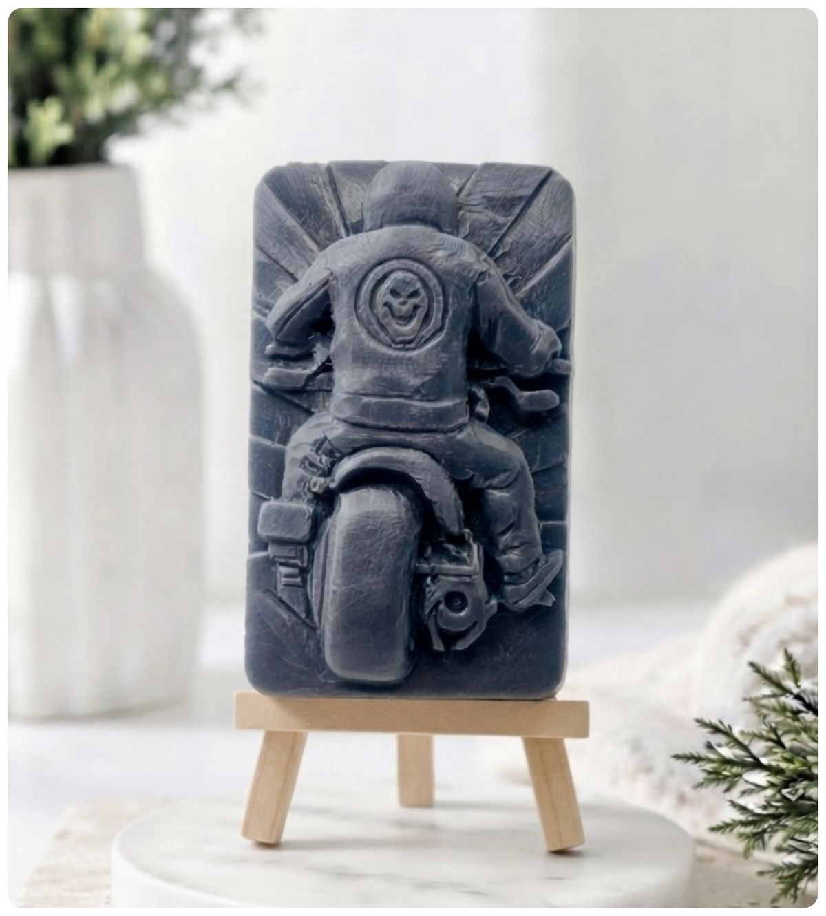 A rectangular handmade vegan soap featuring a detailed high-relief carving of a biker from behind riding a motorcycle, wearing a leather-style jacket with a skull emblem on the back, displayed on a miniature wooden easel.