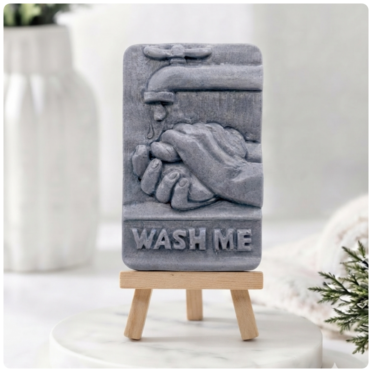 A gray rectangular soap with a raised relief of hands under a faucet and the words “WASH ME,” displayed on a small wooden easel stand against a softly blurred indoor background.