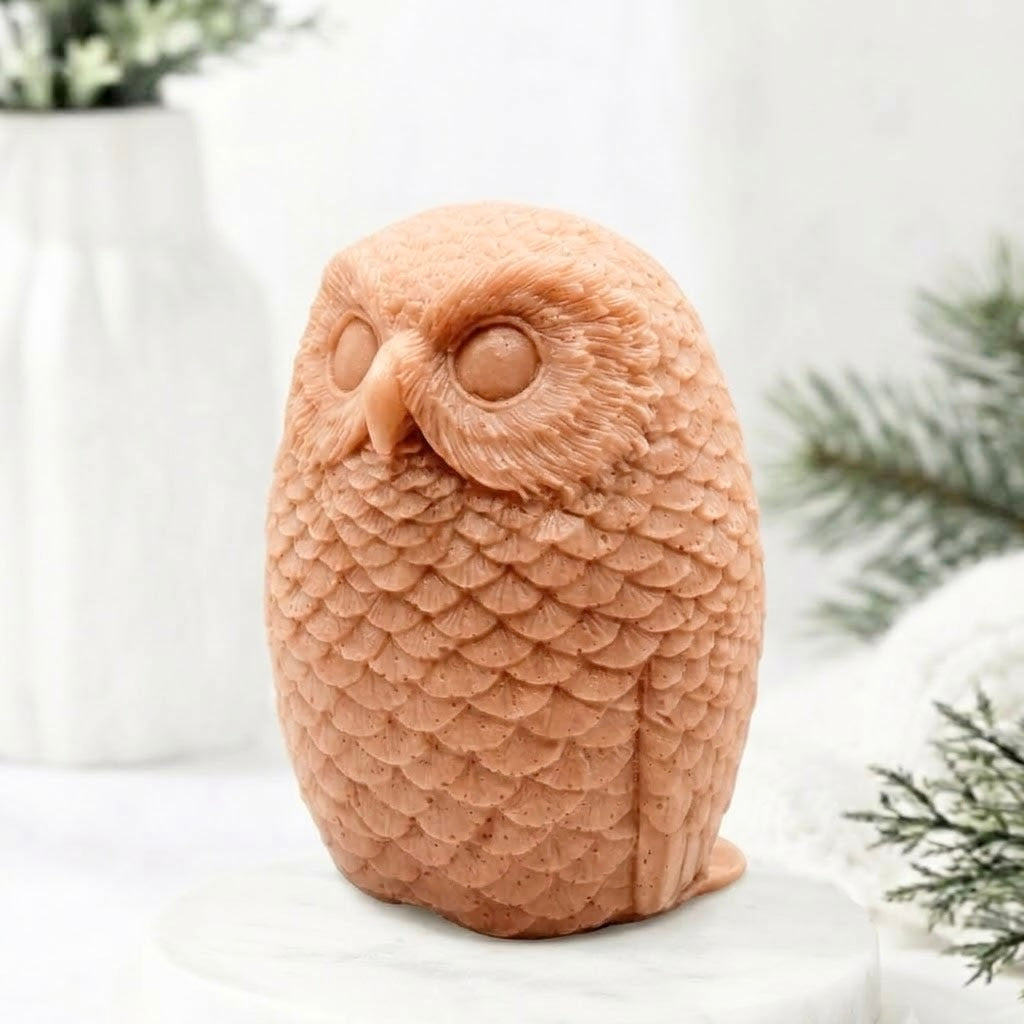 A detailed terracotta coloured handmade vegan soap in the shape of an owl with intricate feather textures, displayed on a light wooden table with white flowers in the background.