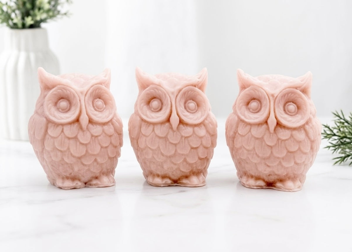 A gift set featuring three soft pink handmade vegan soaps shaped like barn owls , arranged in a row on a light wooden surface with white flowers in the background 