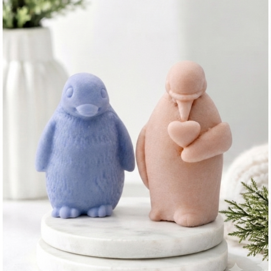 Two cute penguin soaps sitting on a round white marble pedestal. The left penguin is light blue with a textured finish, and the right penguin is a soft peach-pink with a smooth matte finish, holding a small heart.