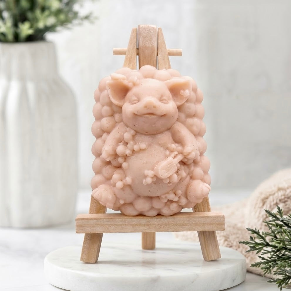 Full-frontal view of a pale pink pig-shaped soap figurine with a bubble-cluster base, sitting on a miniature wooden easel in a clean, neutral environment.