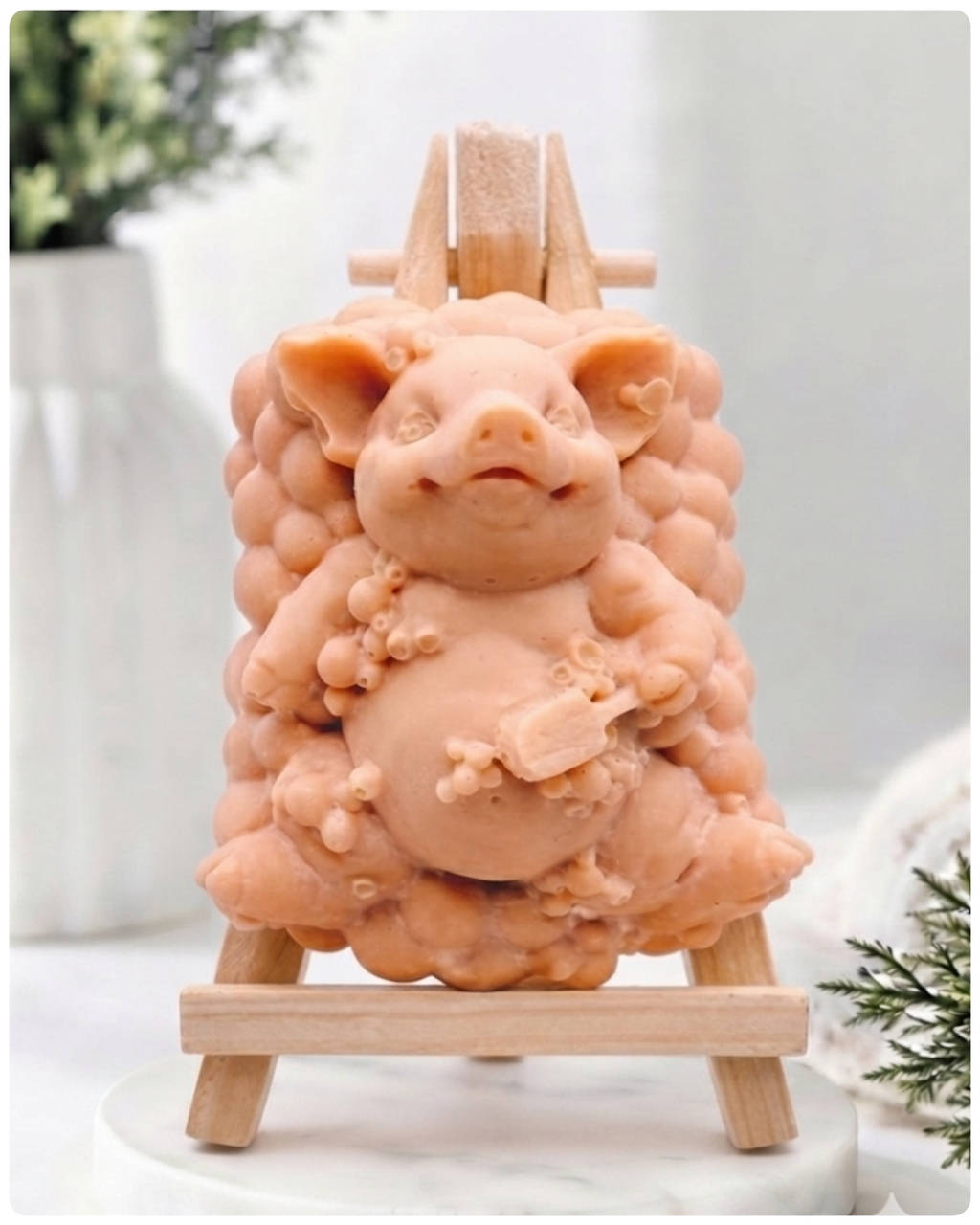 A detailed handmade vegan soap, sculpted as a cheerful pig sitting in a mound of bubbles and holding a scrubbing brush, displayed on a miniature wooden easel against a soft farmhouse window background 