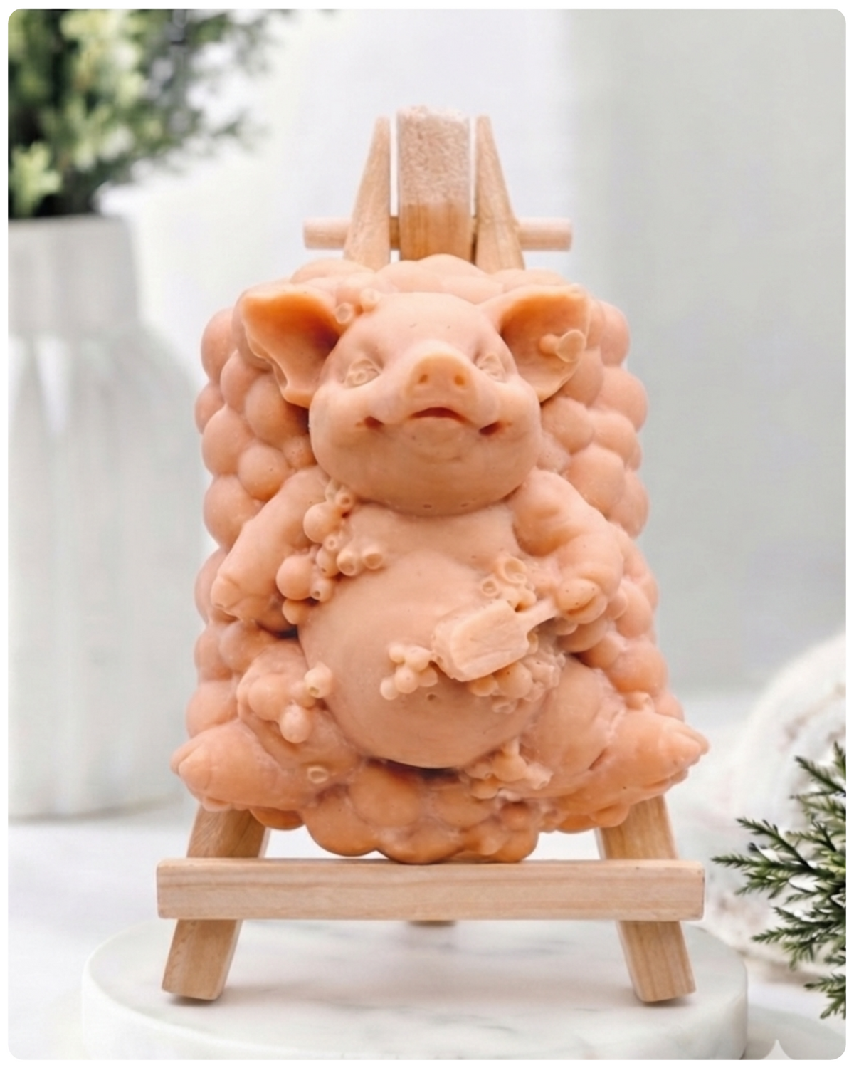 A detailed handmade vegan soap, sculpted as a cheerful pig sitting in a mound of bubbles and holding a scrubbing brush, displayed on a miniature wooden easel against a soft farmhouse window background 