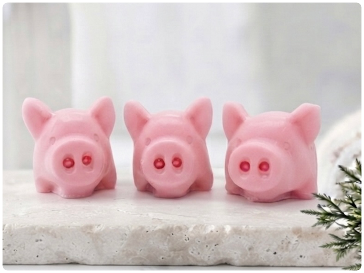 A set of three handmade vegan soaps sculpted into cute pigs with round snouts and floppy ears, displayed with a small decorative bee and ladybird on a white wooden surface.