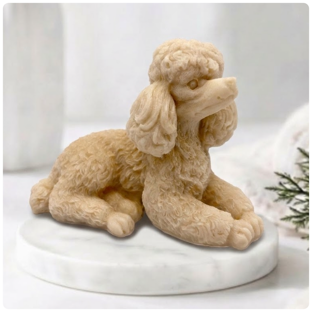 A close-up photograph of a detailed, soap bar shaped like a poodle. The intricate curls of its coat are carved in sharp relief. The poodle-soap is resting on a circular white marble riser. In the soft-focus background are other bathroom accessories and a vase of greenery, all bathed in soft, natural light.