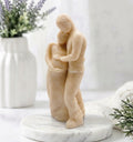 A handmade soft yellow soap sculpture. It depicts a man in pants and a shirt embracing a pregnant woman in a long dress from behind, with his hands over her belly. They are on a round marble stand