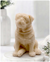 Load image into Gallery viewer, A detailed handmade vegan soap sculpted in the shape of a sitting pug dog with characteristic facial wrinkles and a tilted head, in a natural tone, displayed on a light wooden surface.
