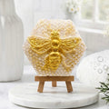 A hexagonal bar of soap, textured like light-gold honeycomb, featuring a prominent, raised golden bee relief on the front. The soap stands upright on a small wooden easel, which is placed on a round, white marble vanity plate.