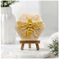 A hexagonal handmade vegan soap featuring a detailed relief of a large queen bee with outstretched wings, highlighted in shimmering gold, set against a textured white honeycomb background and displayed on a miniature wooden easel.