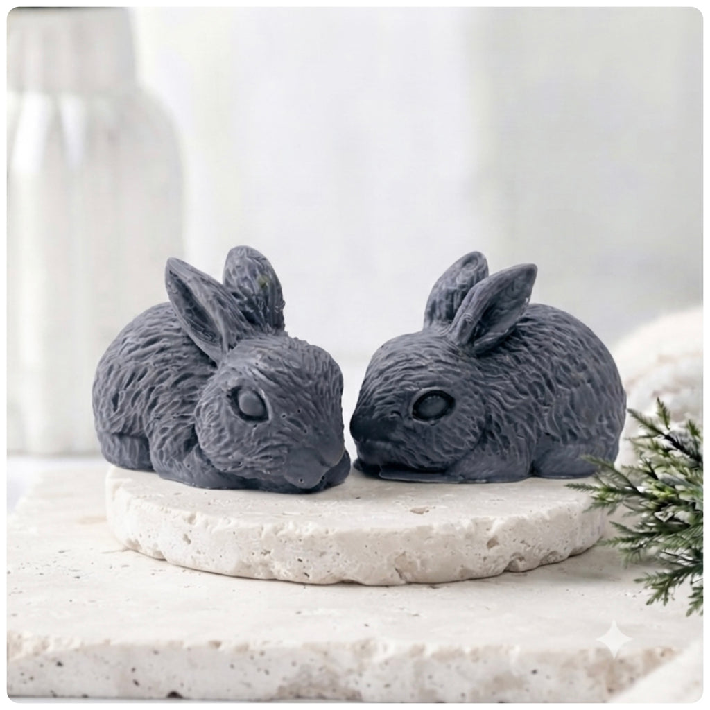 Set of two handmade vegan soaps in the shape of realistic crouching rabbits with detailed fur texture, displayed on a rustic white wooden surface in a bright farmhouse decor setting. Gift boxed. 