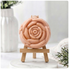 Load image into Gallery viewer, A handmade vegan soap, intricately sculpted in the shape of a large, blooming rose flower with layered petals, displayed on a small wooden easel against a bright win
