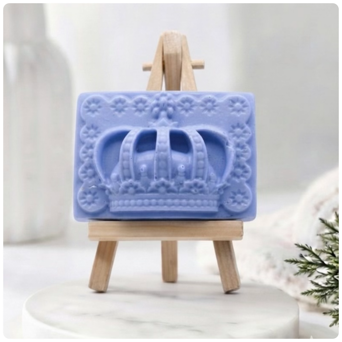 A rectangular handmade vegan soap in a soft tone, featuring a highly detailed relief of a royal crown with floral border accents, displayed on a miniature wooden easel in a bright farmhouse setting.