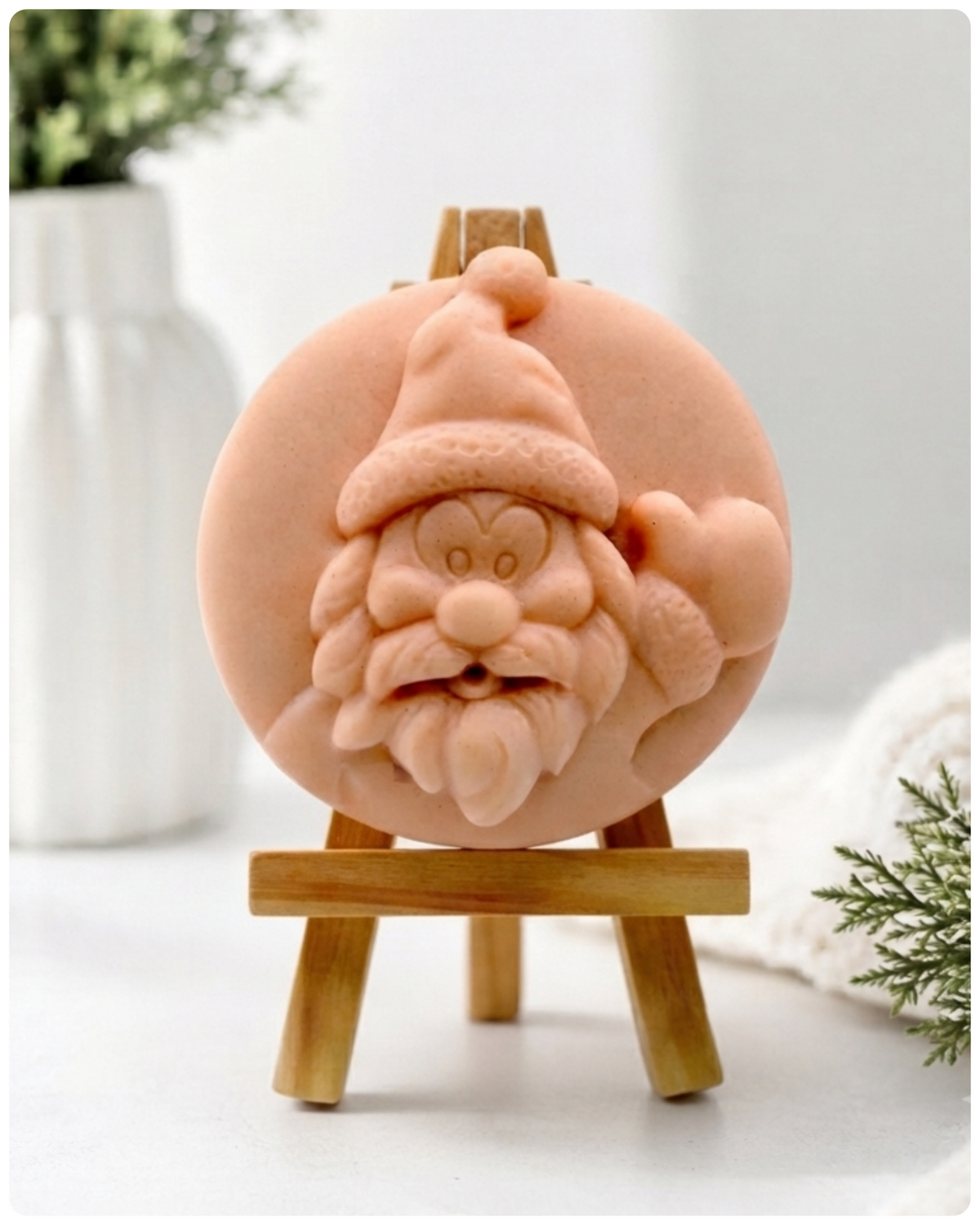 A circular handmade vegan soap featuring a detailed high-relief carving of a jolly Father Christmas waving, with a textured beard and a classic Santa hat, displayed on a miniature wooden easel.