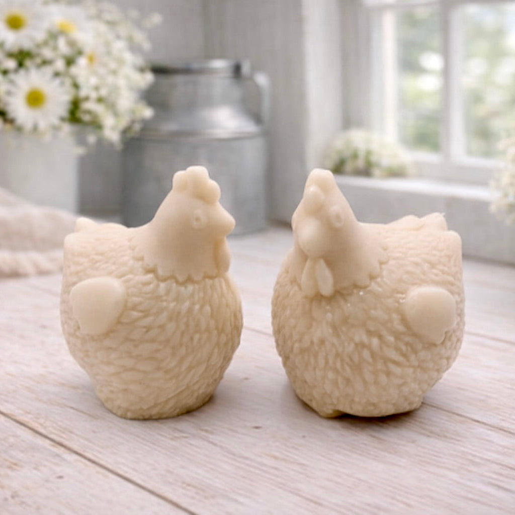 Two handmade vegan soaps in the shape of plump, realistic chicken sculptures with detailed feather textures, displayed on a rustic white wooden surface in a bright farmhouse decor setting.