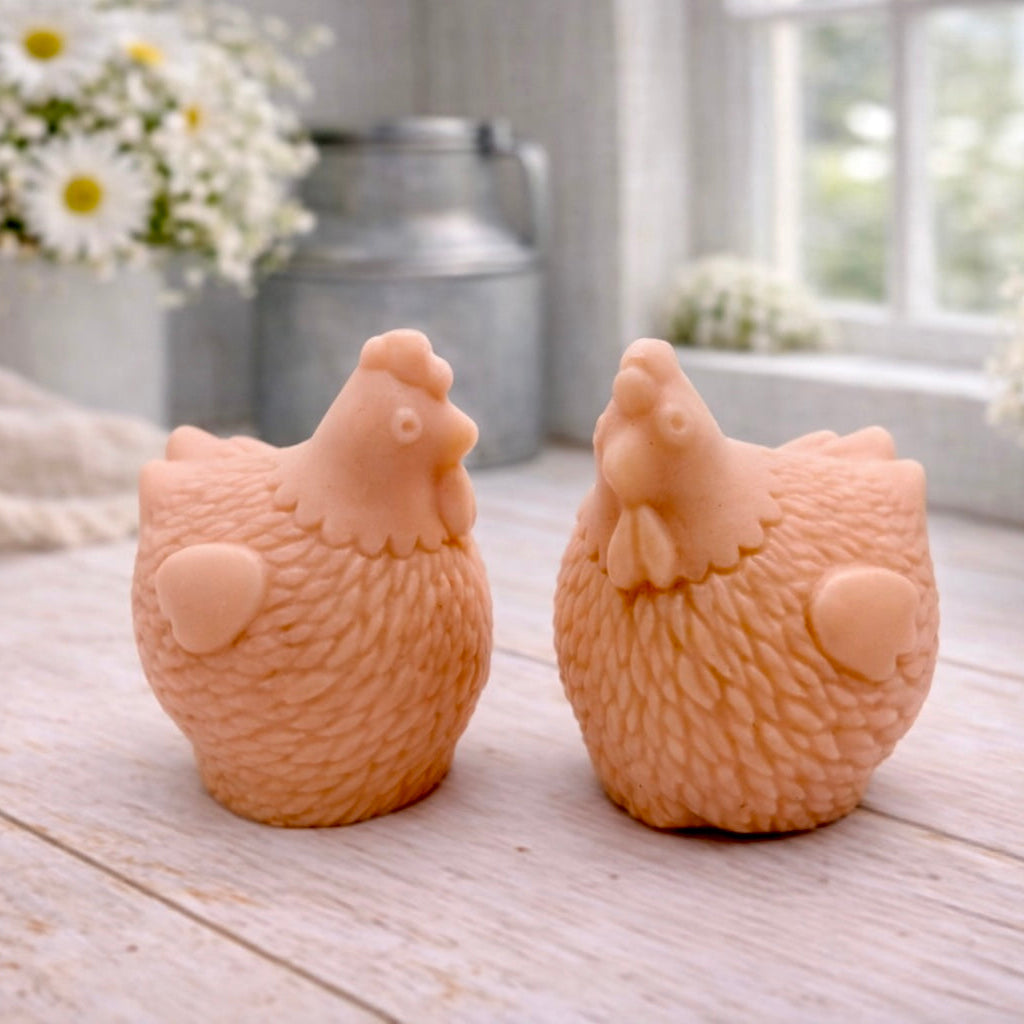 Two handmade vegan soaps in the shape of plump, realistic chicken sculptures with detailed feather textures, displayed on a rustic white wooden surface in a bright farmhouse decor setting.