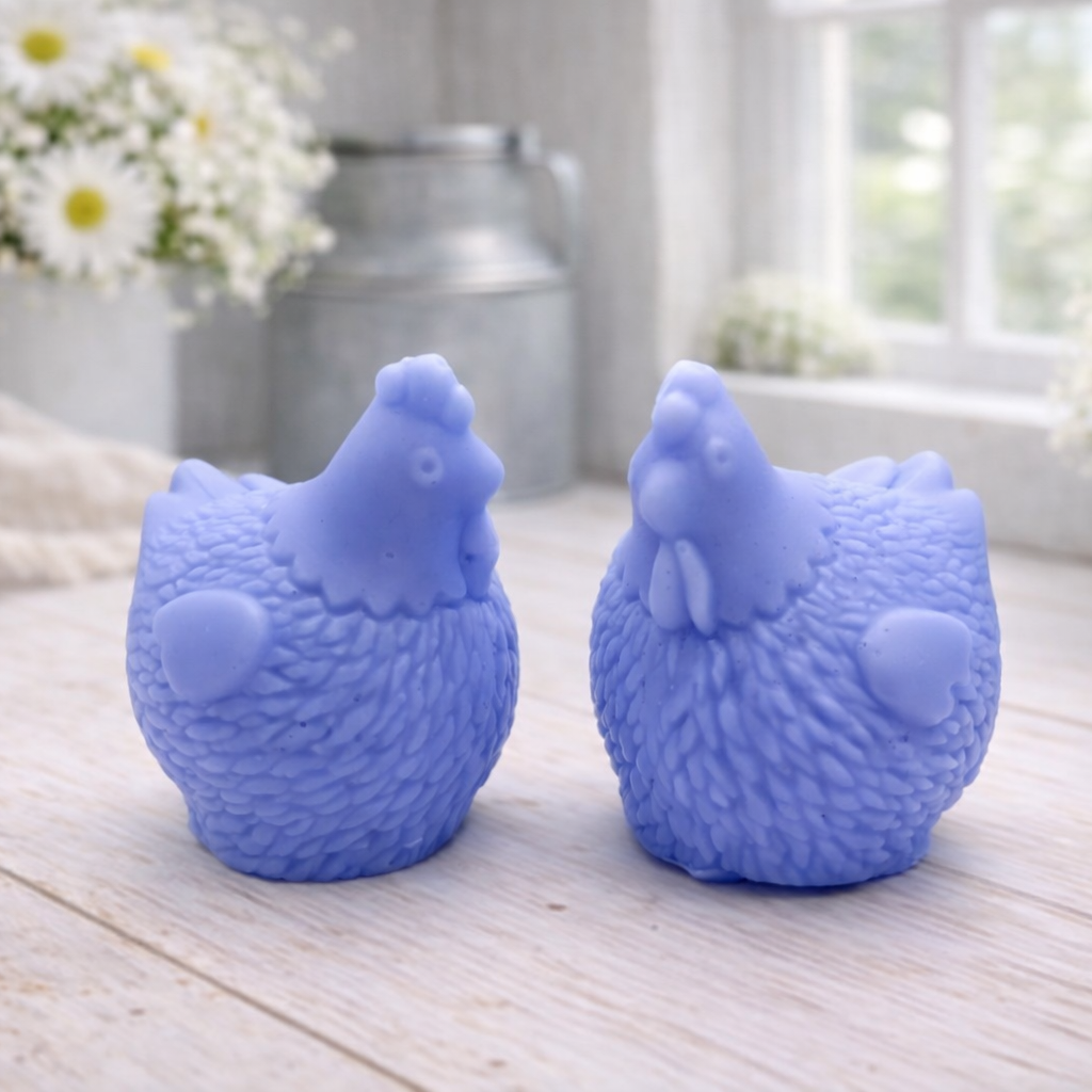 Two handmade vegan soaps in the shape of plump, realistic chicken sculptures with detailed feather textures, displayed on a rustic white wooden surface in a bright farmhouse decor setting.