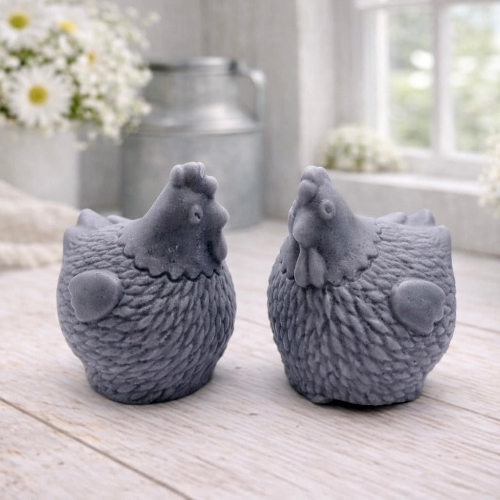Two handmade vegan soaps in the shape of plump, realistic chicken sculptures with detailed feather textures, displayed on a rustic white wooden surface in a bright farmhouse decor setting.