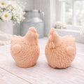 Two handmade vegan soaps in the shape of plump, realistic chicken sculptures with detailed feather textures, displayed on a rustic white wooden surface in a bright farmhouse decor setting.