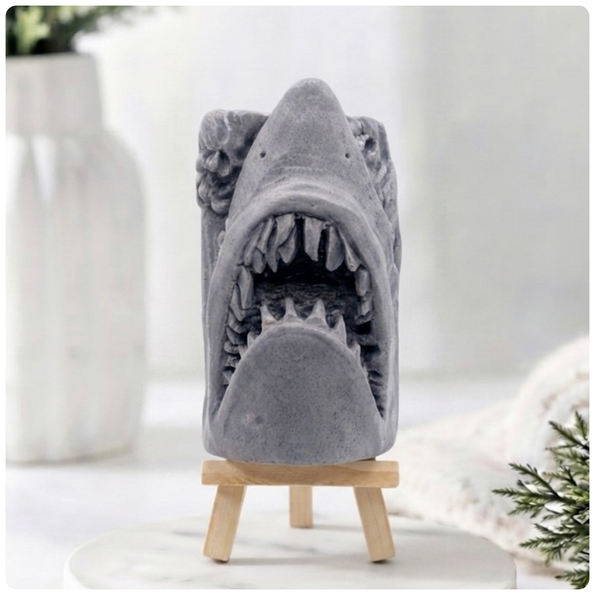 A 3D handmade vegan soap sculpted into a realistic great white shark head with its jaws wide open to reveal multiple rows of sharp teeth, displayed on a miniature wooden easel in a bright farmhouse setting.