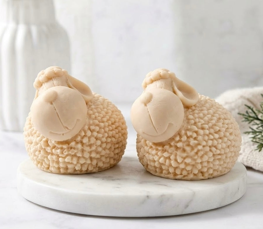 A pair of cute, cream-coloured sheep-shaped soaps with highly textured wool bodies and large ears, sitting on a round white marble stand.