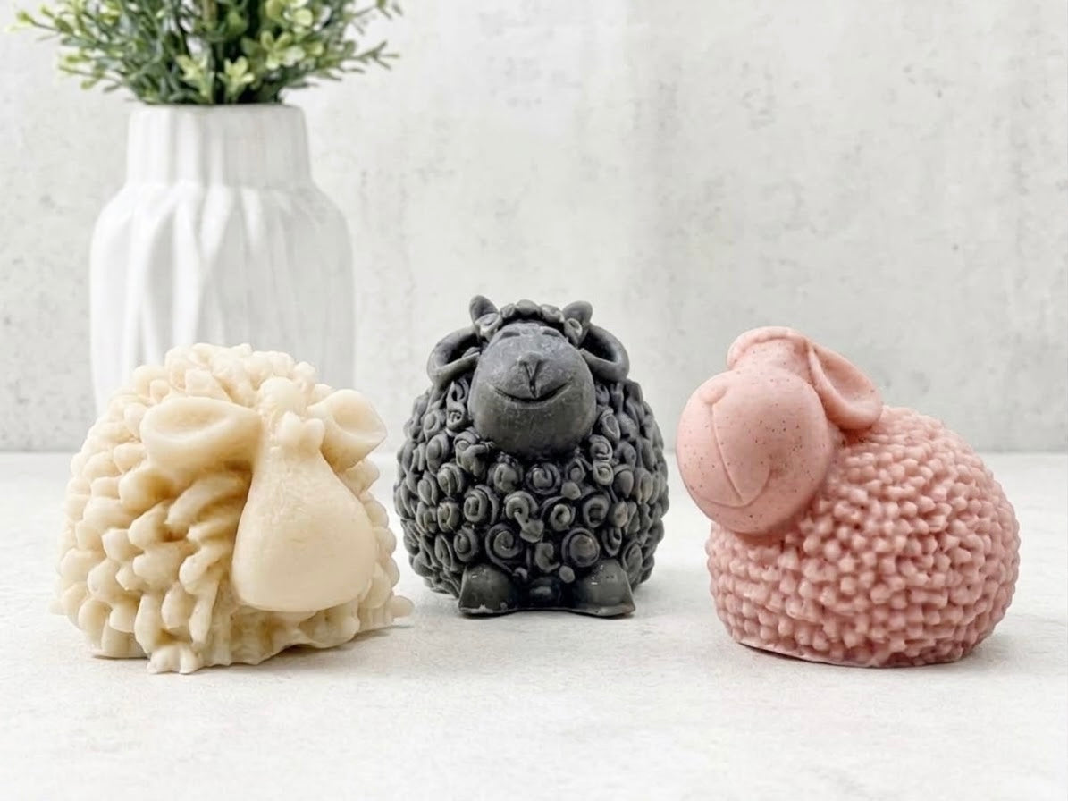 Handmade vegan sheep soap gift set featuring three cute sculptural sheep soaps in cream, charcoal grey, and pink on a light surface with a white vase and greenery background.