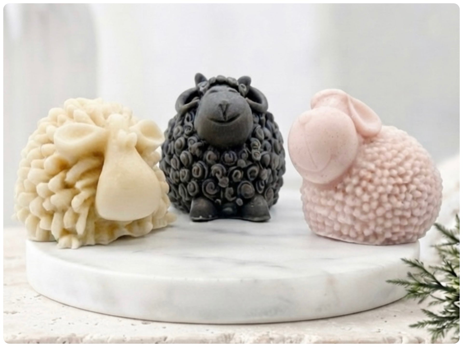 A set of three handmade vegan soaps in different sheep styles, including a dark grey smiling sheep and two lambs with detailed woolly textures, displayed on a rustic wooden surface with lavender and flowers.