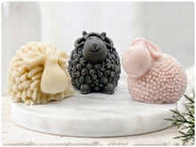 Load image into Gallery viewer, A set of three handmade vegan soaps in different sheep styles, including a dark grey smiling sheep and two lambs with detailed woolly textures, displayed on a rustic wooden surface with lavender and flowers.

