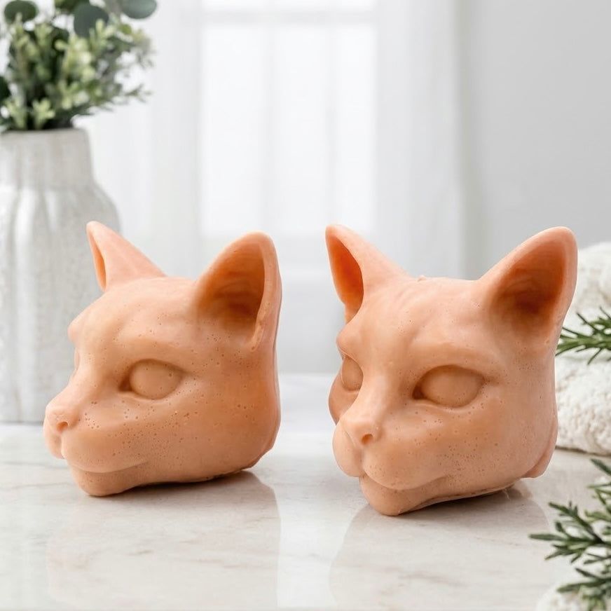 Two terracotta Siamese cat head vegan soap bars shown in side-profile and three-quarter views, resting on white marble against a soft, sunlit bathroom background with greenery.