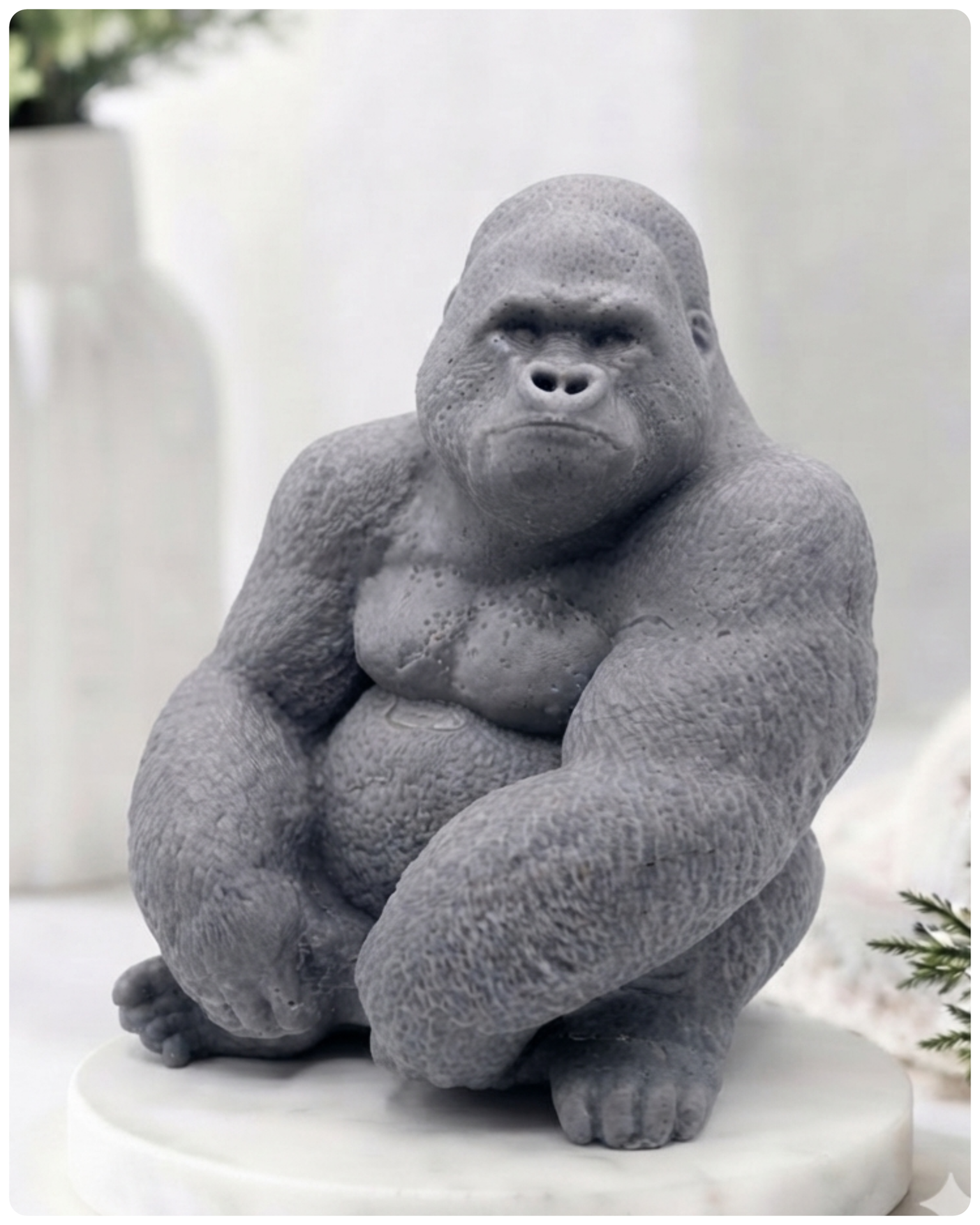 A large gray gorilla handmade soap sitting on a white marble base, photographed in a softly styled indoor setting with blurred decor in the background.