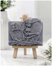 Load image into Gallery viewer, A handmade vegan soap depicting a skeleton resting between two R.I.P. gravestones, displayed on a small wooden easel.
