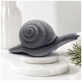 A highly detailed handmade vegan soap in a slate-grey tone, sculpted in the shape of a realistic snail with a prominent spiralled shell and a textured, leathery body, displayed on a light wooden surface.