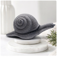 Load image into Gallery viewer, A highly detailed handmade vegan soap in a slate-grey tone, sculpted in the shape of a realistic snail with a prominent spiralled shell and a textured, leathery body, displayed on a light wooden surface.
