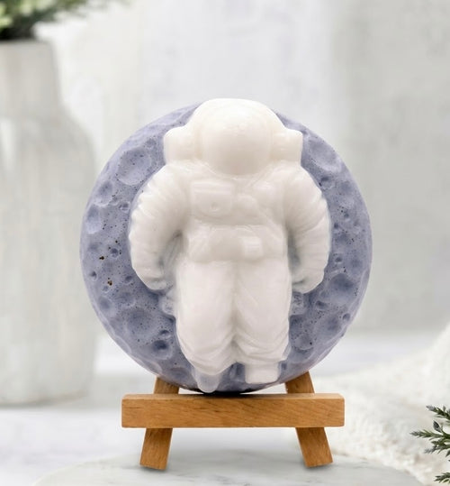 A detailed, close-up photograph of a round, artisan vegan soap bar. The white relief carving of a detailed astronaut stands out against a blue, speckled backdrop that looks like the surface of a moon. The soap rests on a miniature wooden artist's easel.