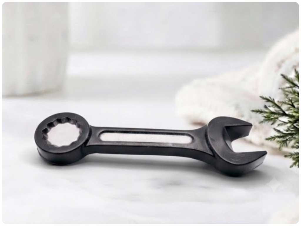 A 3D handmade vegan soap sculpted in the shape of a realistic spanner or wrench tool, finished in a sleek charcoal black with silver-painted detailing on the handle and head, lying on a light wood surface.