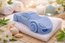 Load image into Gallery viewer, Blue car-shaped vegan soap on a white towel with flowers and candles in the background
