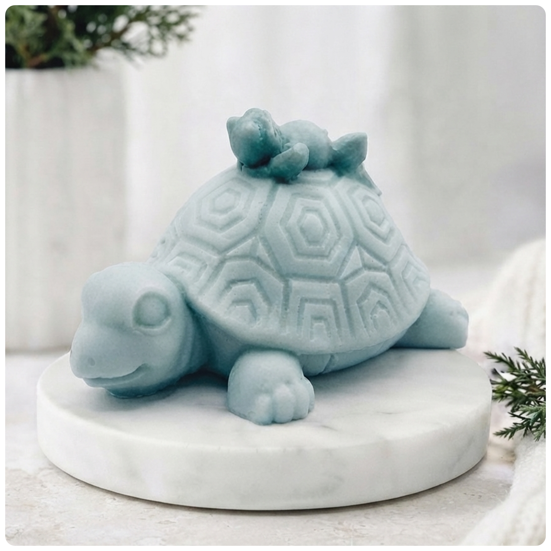 A handmade vegan soap in a soft mint green tone, sculpted into a large friendly turtle with a geometric shell pattern and a small, cute frog sleeping peacefully on its back.