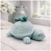 Load image into Gallery viewer, A handmade vegan soap in a soft mint green tone, sculpted into a large friendly turtle with a geometric shell pattern and a small, cute frog sleeping peacefully on its back.
