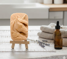Load image into Gallery viewer, handmade vegan soap featuring a detailed unicorn head relief with a spiralled horn and flowing curly mane, displayed on a small wooden easel in a bright farmhouse decor setting.
