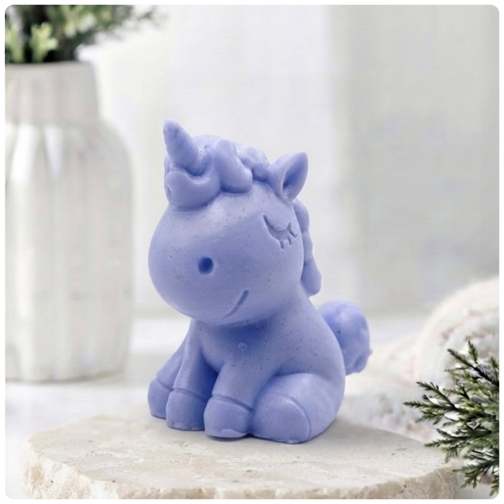 A handmade vegan soap sculpted into a cute sitting unicorn with a spiralled horn, a smiling expression with a closed winking eye, and a bubbly mane and tail, displayed on a light wooden surface in a bright farmhouse setting.