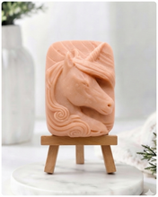 Load image into Gallery viewer, handmade vegan soap featuring a detailed unicorn head relief with a spiralled horn and flowing curly mane, displayed on a small wooden easel in a bright farmhouse decor setting.
