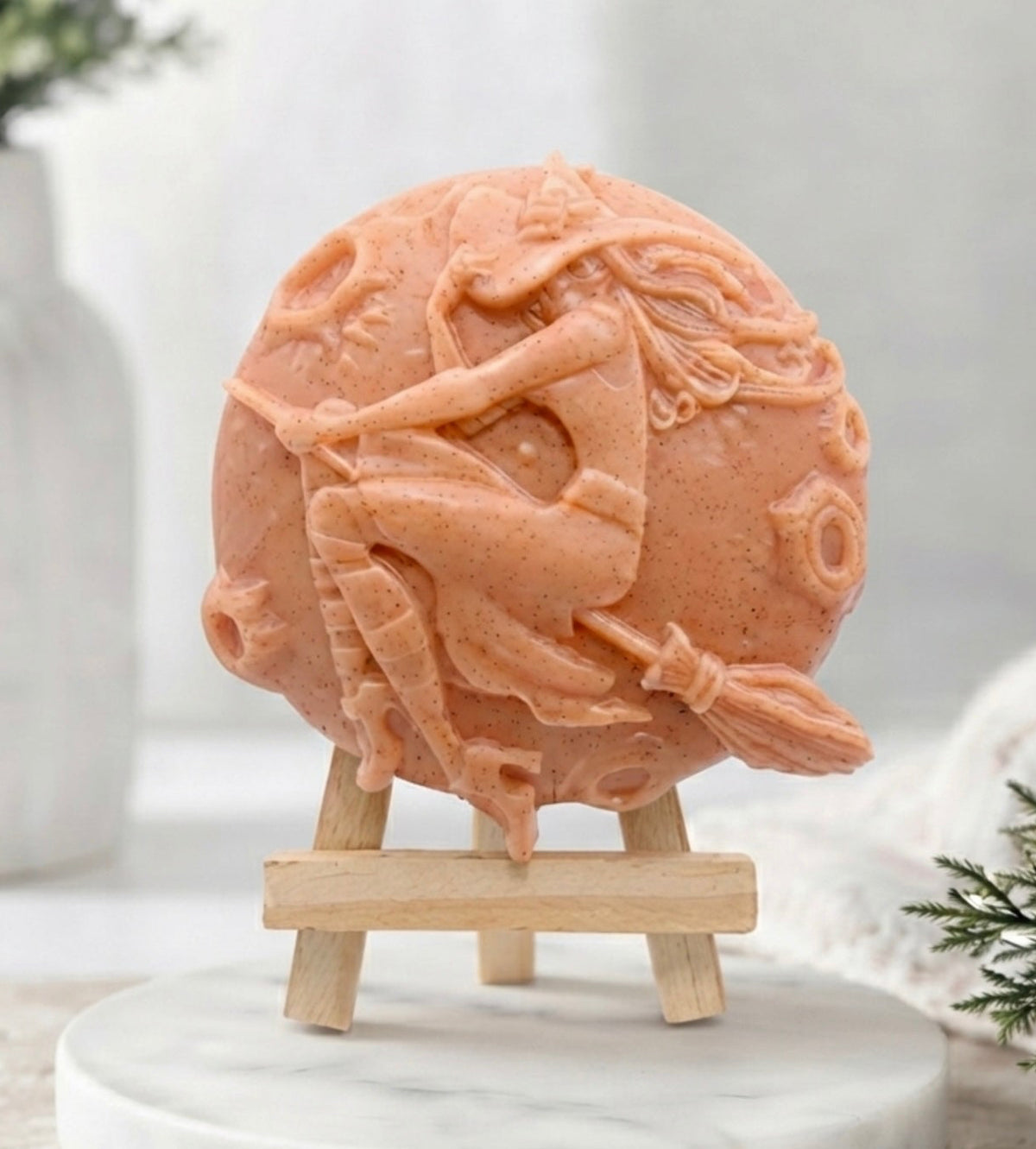 A round red French clay vegan soap with a detailed carving of a witch riding a broomstick against a full moon. The soap is displayed upright on a small wooden easel stand. 