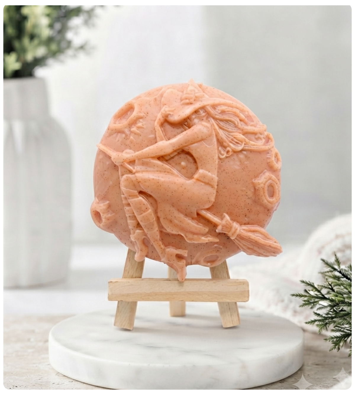 A circular handmade vegan soap featuring a detailed high-relief carving of a witch wearing a pointed hat, flying on a broomstick across a cratered moon, displayed on a miniature wooden easel.
