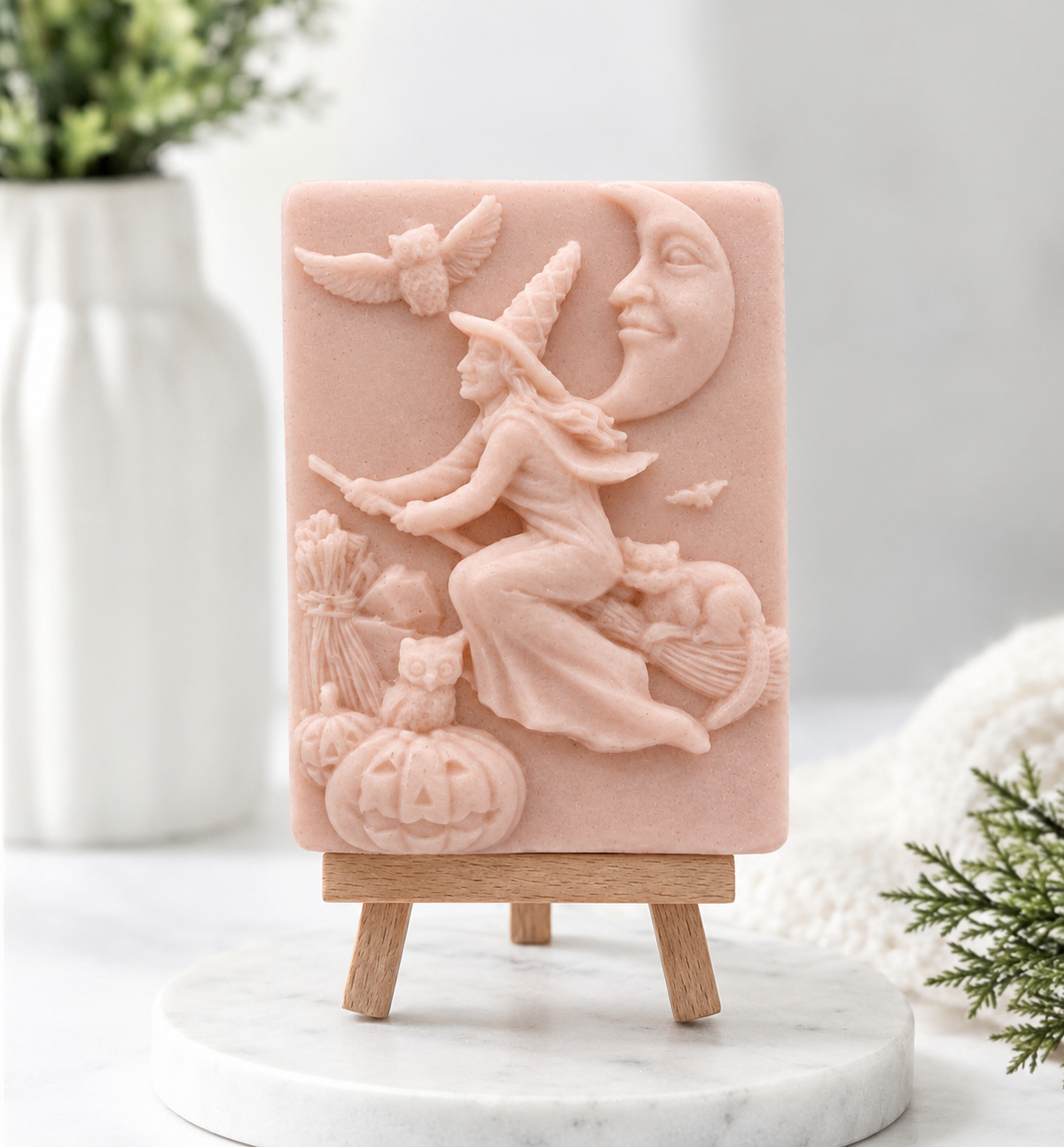 A rectangular pale pink decorative soap with a raised Halloween scene featuring a witch riding a broomstick beside a crescent moon, with pumpkins, birds, and bats, displayed on a small wooden stand.