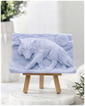 A rectangular handmade vegan soap, featuring a detailed relief carving of a wolf in mid-stride across a rugged landscape with a large full moon in the background, displayed on a miniature wooden easel.