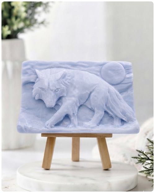A rectangular handmade vegan soap, featuring a detailed relief carving of a wolf in mid-stride across a rugged landscape with a large full moon in the background, displayed on a miniature wooden easel.
