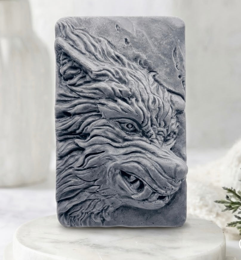 A rectangular charcoal grey vegan soap featuring a highly detailed, sculpted wolf head in relief, displayed on a round white marble pedestal.