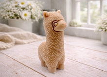 Load image into Gallery viewer, handmade vegan soap in the shape of a realistic standing alpaca sculpture, featuring detailed curly wool texture and a smiling face, displayed on a rustic white wooden surface in a bright farmhouse decor setting.
